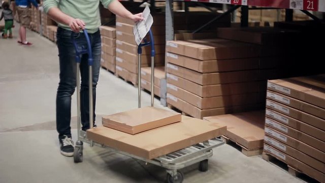 Waist-deep view of a man checking his list and pushing a trolley with boxes on it in a warehouse