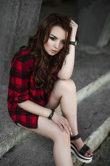 Naklejka premium Beautiful young hipster girl posing and smiling near urban wall background in red plaid shirt, shorts, outdoors summer portrait.