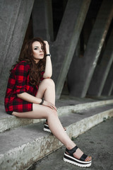 Beautiful young hipster girl posing and smiling near urban wall background in red plaid shirt, shorts, outdoors summer portrait.