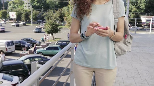 Slender young girl uses a pink mobile phone on city background in slow motion. Woman in a T-shirt with a backpack in the city in the summer.