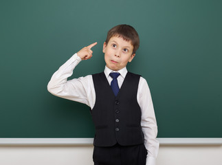 school student boy posing at the clean blackboard, grimacing and emotions, dressed in a black suit, education concept, studio photo