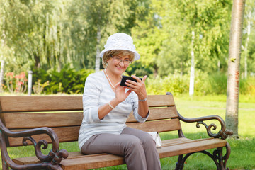 Attractive mature woman sitting on a bench holding and using a smartphone mobile device with touch screen, outdoors.