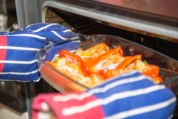 Cooked stuffed red peppers with eggs, ham and croutons in an oven proof serving dish being taken from an oven with red and blue striped oven gloves.