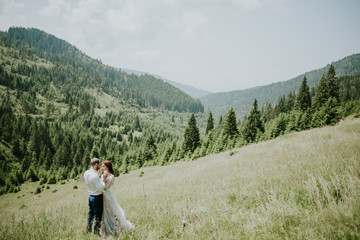 Naklejka premium Vintage wedding photography of a young hipster couple curly bride posing at sunset in the forest and mountains dressed in a suite and white dress