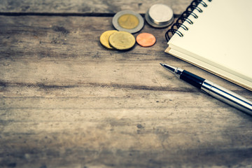 close up pen, notepad and coins on wooden background with copy space. Financial planning concept. Selective focus with vintage tone photo.