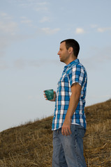 Man standing and looking out onto a hill side holding a cup in his hand wearing a plaid shirt.