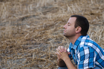 Man crouching down and praying with his hands clasped on a hill side wearing a plaid shirt.