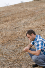 Man crouching down and praying with his hands clasped on a hill side wearing a plaid shirt.