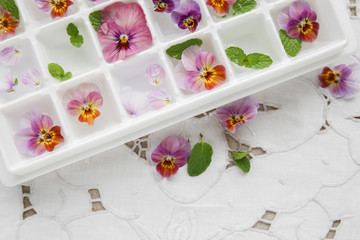 Obraz na płótnie Canvas Edible flowers and mint in ice cubes tray on white vintage linen
