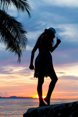 Young woman dancing on the rocks by the sea against the backdrop of palm trees and sunset
