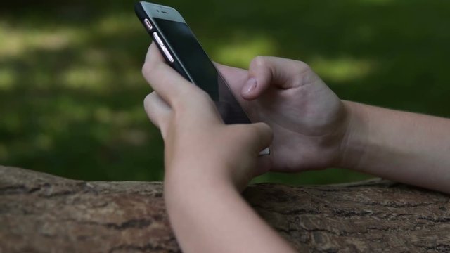 Close-up shot of a teengaer girl typing message on smart phone leaning on the tree in the park