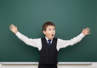 school student boy posing at the clean blackboard and open arms, grimacing and emotions, dressed in a black suit, education concept, studio photo