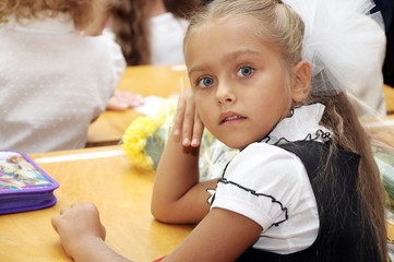 Fototapeta premium Beautiful girl first grader in a school uniform sitting at a desk in a classroom. Schoolgirl with folded hands sitting at school desk at lesson. Girl in school uniform, Knowledge Day 