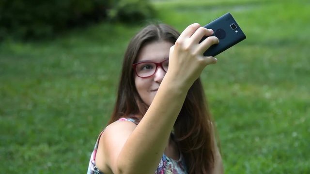 Brunette girl teenager in glasses sitting on the grass in park and taking selfie with cellphone