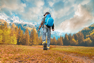 Little boy with backpack walking in nature. Autumn time, cloudy sky and snowly peak of mountains