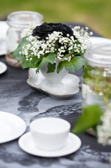 Modestly decorated wedding table. Gothic romance, garden wedding. White cups standing on a black tablecloth with handmade white pattern. A bouquet of white and black flowers