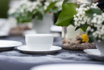 Modestly decorated wedding table. Gothic romance, garden wedding. White cups standing on a black tablecloth with handmade white pattern. Clean light, white plates, handcrafted decoration, black roses.