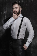 Bearded man in a white shirt and suspenders standing near dark wall. He is attractive, strong, courageous and serious. He strokes his thick dark beard.