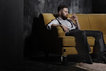 A handsome brutal bearded man sitting on an orange couch. He is holding an glass with some drink. Half empty bottle standing on the floor. Dark room, old carpet, deep shadows.