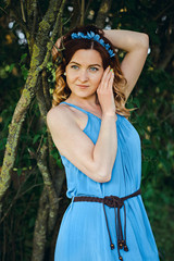Old fashion portrait of a beautiful elegant red-haired woman dressed in a blue dress and a wreath of flowers posing in a windy Sunny day in a summer outfit, in nature. European city