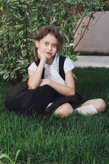 Cute elementary schoolgirl in uniform sitting with backpack on grass. Schoolgirl in a black dress and a white blouse and gaiters