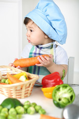 Little boy with serious face in kitchen apron and cap holding carrot
