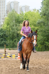 Fototapeta premium Happy woman with curly hair riding a horse in park near the apartment complex