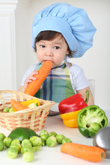 Little boy with serious face in kitchen apron and cap eating carrot