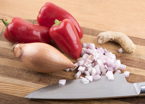 Image of red peppers, onions and ginger on a wooden chopping board with a knife.
