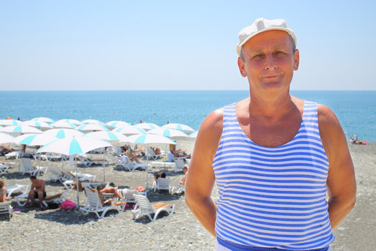 Portrait of elderly man in a striped vest and a white cap on the beach near the sea - Powered by Adobe