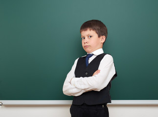 school student boy posing at the clean blackboard, grimacing and emotions, dressed in a black suit, education concept, studio photo