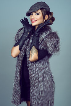 Portrait of a beautiful glam smiling model with provocative make up wearing silver fox jacket, long leather gloves and peaked cap. Natural furs concept. Studio shot.
