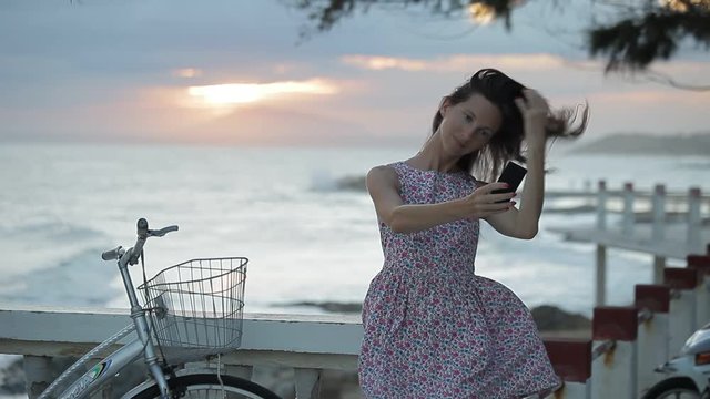 Girl in summer dress is making selfie on the embankment near a beach at sunset.