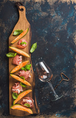 Italian antipasti snack for wine. Prosciutto ham with cantaloupe melon, basil leaves and wine glass on wooden serving board over grunge dark plywood background. Top view, copy space