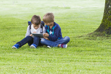 Naklejka na ściany i meble Little boys sitting on the grass in a park and using tablet PC. Technology, lifestyle, education, people concept
