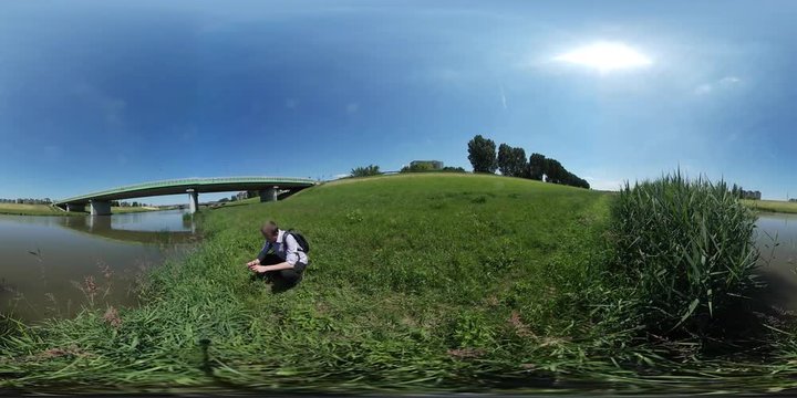 360Vr Video Tourist Sits Down on a Grass Landscape Taking Photo of Green Meadow by Small River Bridge Through River Trees Wilderness in Sunny Summer Day