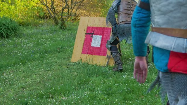 A knight is having a difficulty getting a hatchet out of a target. He is going to do another round of throwing small hatchets. Wide-angle shot.
