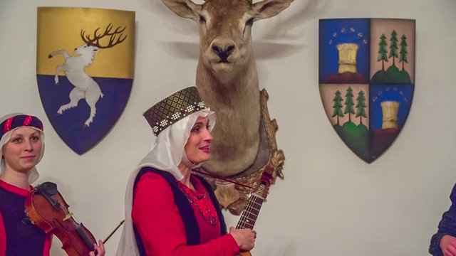 A princess is saying something abou a stuff deer's head which is hanging on a wall. She is holding a tambura in her hands. Close-up shot.
