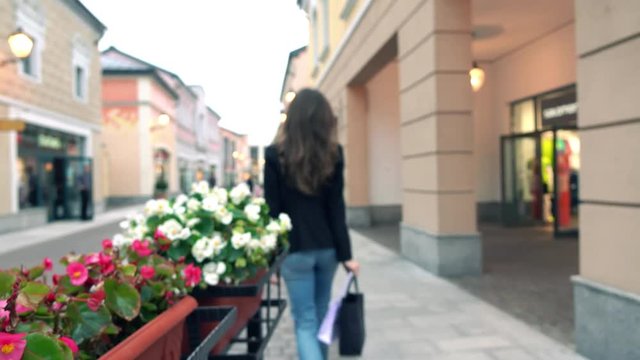 Blurred slim brunette girl in jeans walks with her purchasings in bags near outlet stores. 4K bokeh background video
