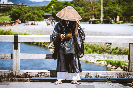 Japanese Man Wearing Traditional Costume And Big Hat Hiding His Face Stands On The Street Of Kyoto With A Wooden Bow