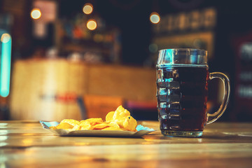 A fresh glass of cold dark beer and potato chips on the wooden bar counter in pub with free space...