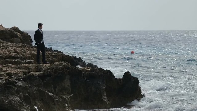 Handsome man in suit standing on the rocky shore and watches the sea waves
