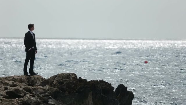 Handsome man in suit standing on the rocky shore and thoughtfully watches the sea waves on back view