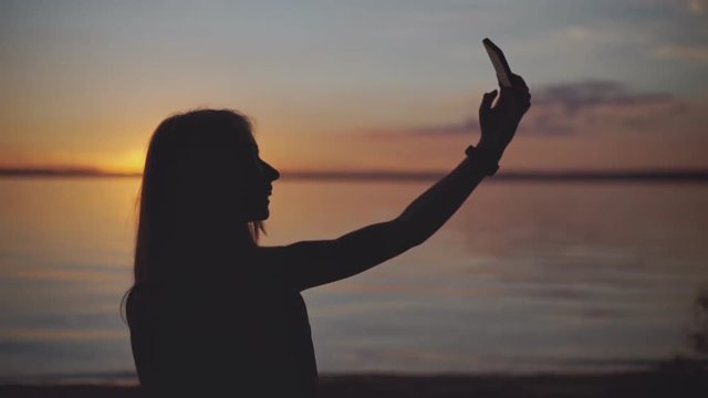 Silhouette of young girl taking photo selfie on the beach at the sunset