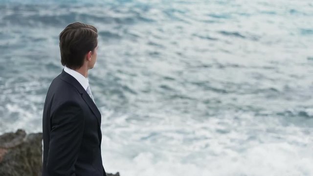 Handsome businessman in suit standing on the rocky shore and thoughtfully watches the sea waves on back view