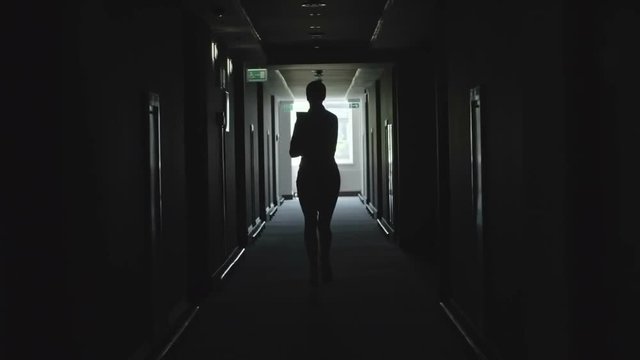 Rear view of silhouette of businesswoman running alone through dark corridor in office building and holding documents 