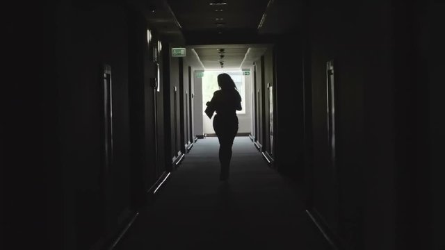 Backlit silhouette of woman with long fly away hair running alone through dark corridor in office building and holding file 