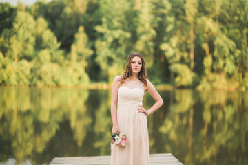 Gorgeous bride in elegant dress holding bouquet posing near forest and lake