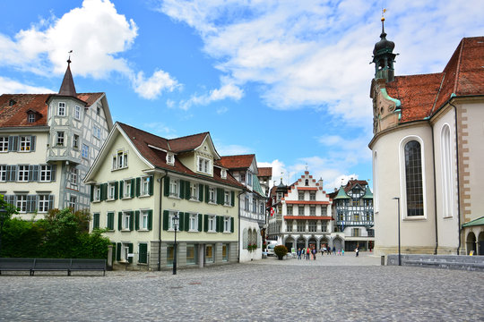 A picturesque view of Gallusplatz in St. Gallen, Switzerland, featuring traditional houses with green shutters and the historic cathedral. The charming square captures the essence of Swiss urban