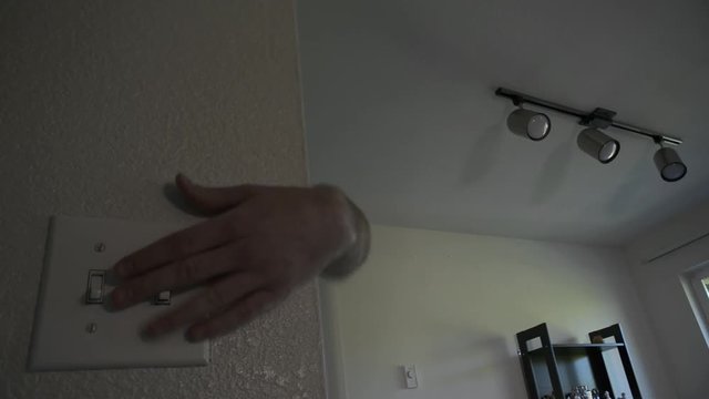 A hand reaching around the corner of a wall to flick switches, turning on and off the overhead track lights. Both lights and switch in focus.